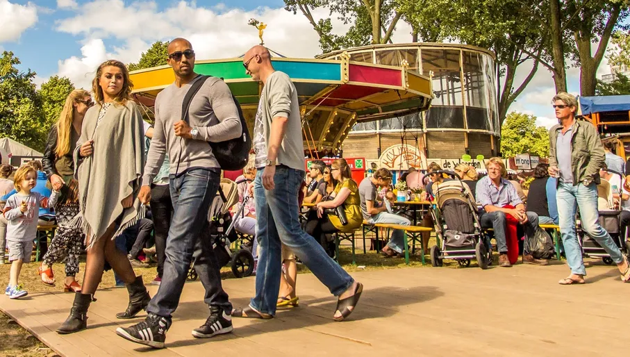 Diverse festival-goers enjoy a vibrant outdoor summer event in Amsterdam, strolling past a colorful carousel and relaxing at picnic tables under a bright sky.
