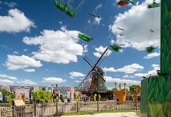 Holland-Park delights with a traditional Dutch windmill, blurred green swing rides in motion under a bright blue sky, colorful themed buildings, and a distinctive yellow clog ride, embodying a cheerful amusement park experience.