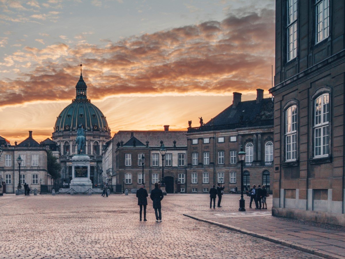 Visitors stroll through Amalienborg Palace Square in Copenhagen, admiring the equestrian statue of King Frederik V and the majestic Frederik's Church dome against a glowing sunset.