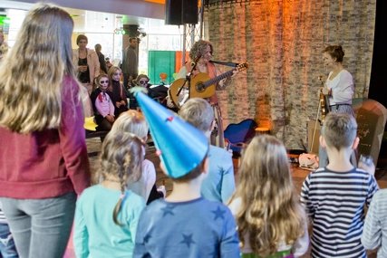 Children and adults eagerly watch a live musical performance on a theatre stage at FEZ Berlin, where two artists entertain with guitar and a whimsical tombstone prop.