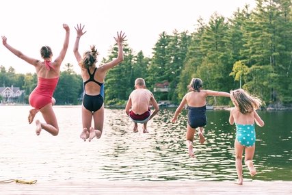 Five happy young people in swimsuits leap from a wooden dock into a calm lake, surrounded by a dense treelined shore, highlighting a refreshing summer swimming spot.