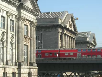 Vibrant red train travels on an elevated track, passing ornate classical buildings of Berlin's historic Museumsinsel bathed in sunlight.