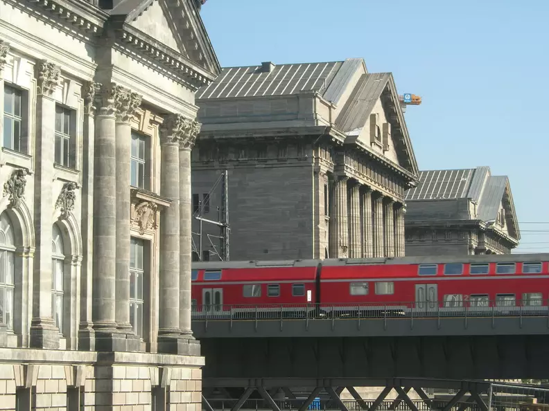 A bright red train crosses a bridge, with the grand classical stone buildings of Berlin's Museumsinsel visible under a clear blue sky.