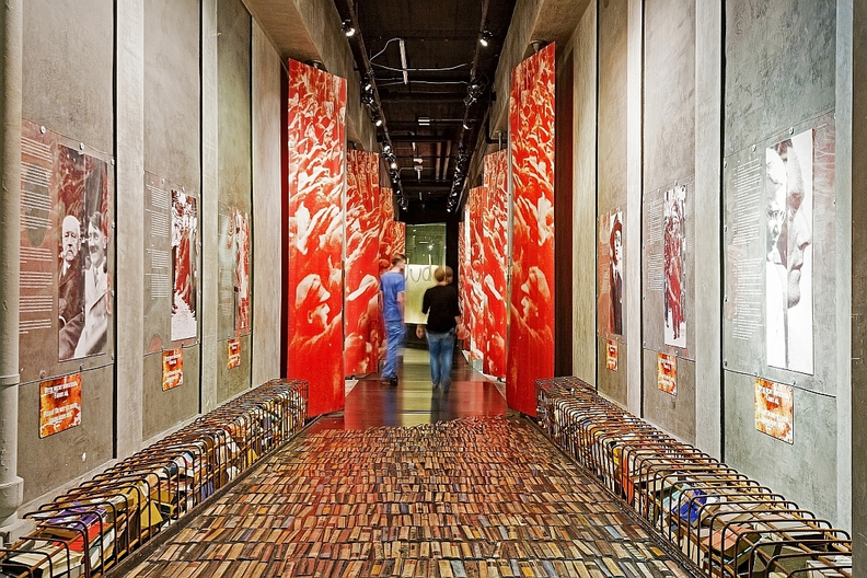 An immersive museum exhibition on Berlin's National Socialist history, featuring a corridor paved with countless books symbolizing book burning, flanked by towering red banners depicting mass rallies and historical photographs on concrete walls.