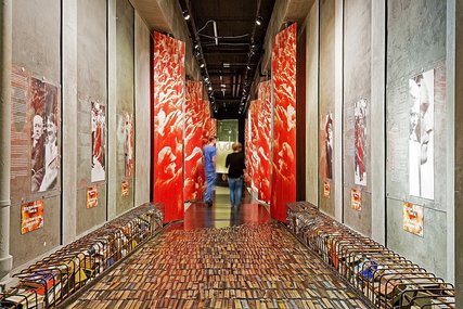 Somber museum exhibit corridor showcasing a floor paved with books, towering red panels with abstract designs, and historical posters on concrete walls, powerfully evoking Berlin's Nazi-era book burnings.