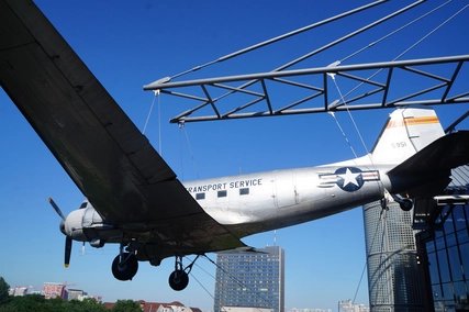 Historic silver Douglas C-47 Rosinenbomber aircraft, a symbol of the Berlin Airlift, prominently suspended outdoors at the Deutsches Technikmuseum Berlin against a clear blue sky and modern city skyline.
