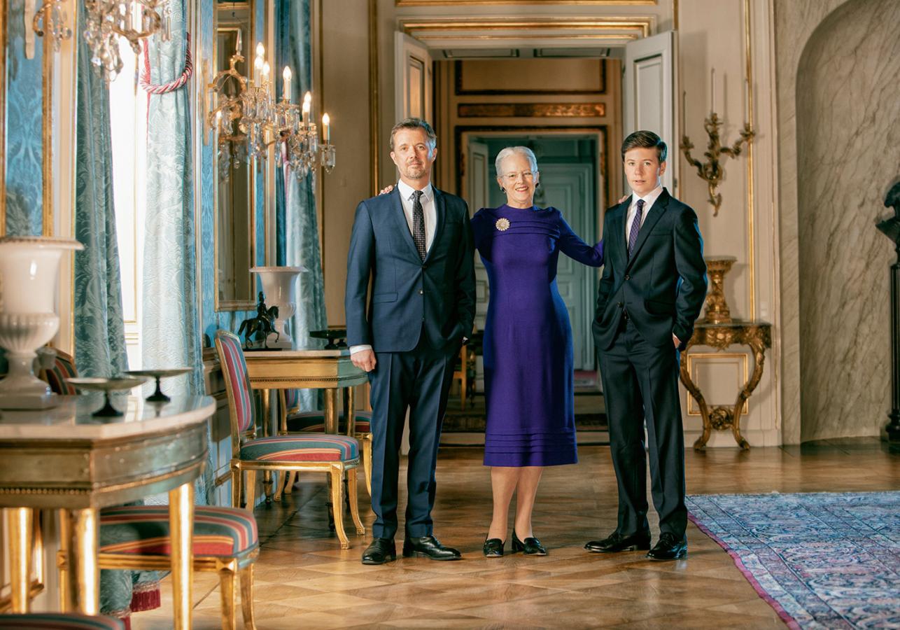 Queen Margrethe II of Denmark in a vibrant purple dress stands formally with Crown Prince Frederik and Prince Christian in a grand, richly decorated Copenhagen royal palace hall.