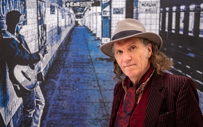 A confident artist in a fedora and velvet jacket stands before a vibrant blue street art mural depicting a subway station with a guitarist figure, embodying Paris's urban art scene.