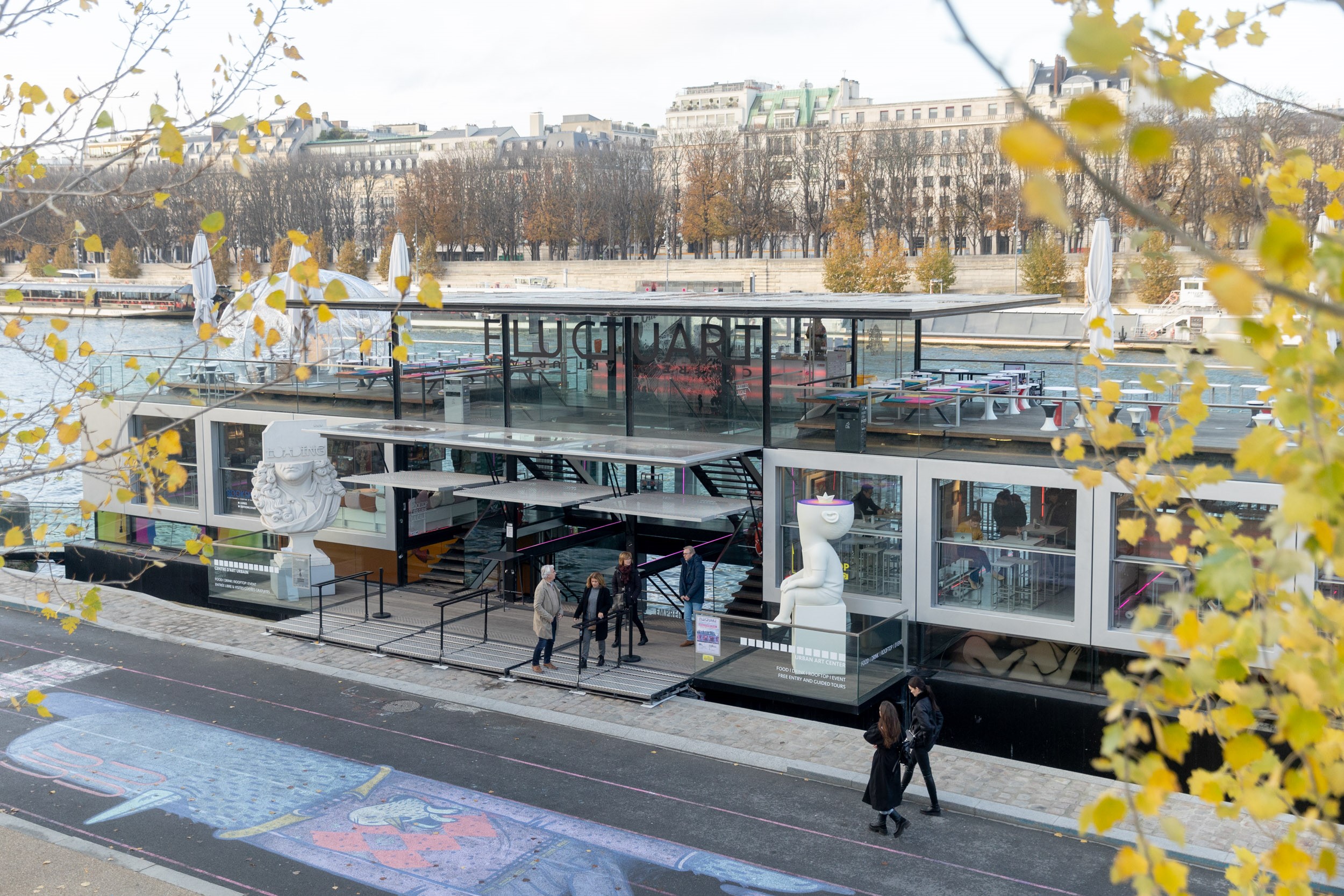 Fluctuart, a modern glass urban art center boat, is moored on the Seine River in autumn Paris, featuring large contemporary sculptures, a lively quayside with a colorful street art mural, and distant city buildings.