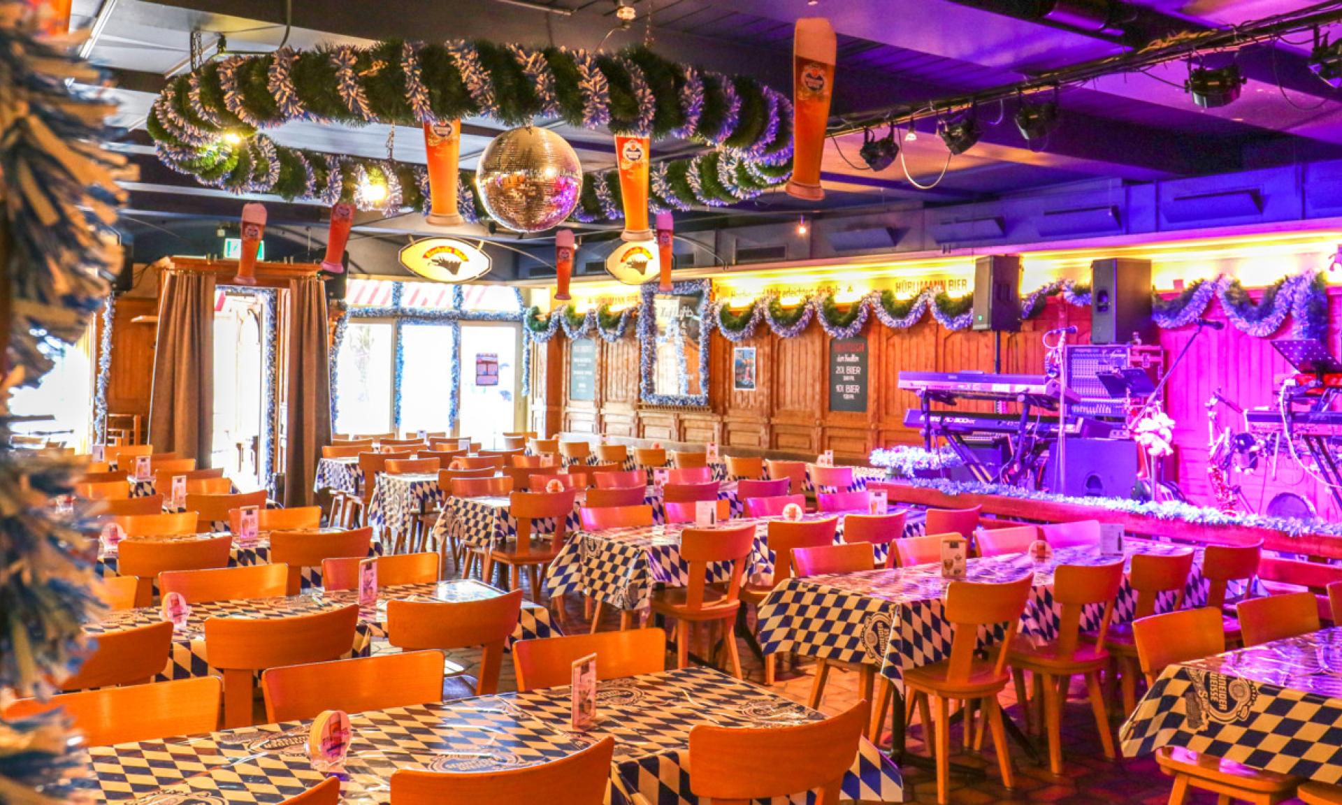 Vibrant Bierhalle Wolf interior with rows of tables featuring blue and white checkered tablecloths, wooden chairs, festive green garlands, a disco ball, and a lively, purple-lit stage for live music.