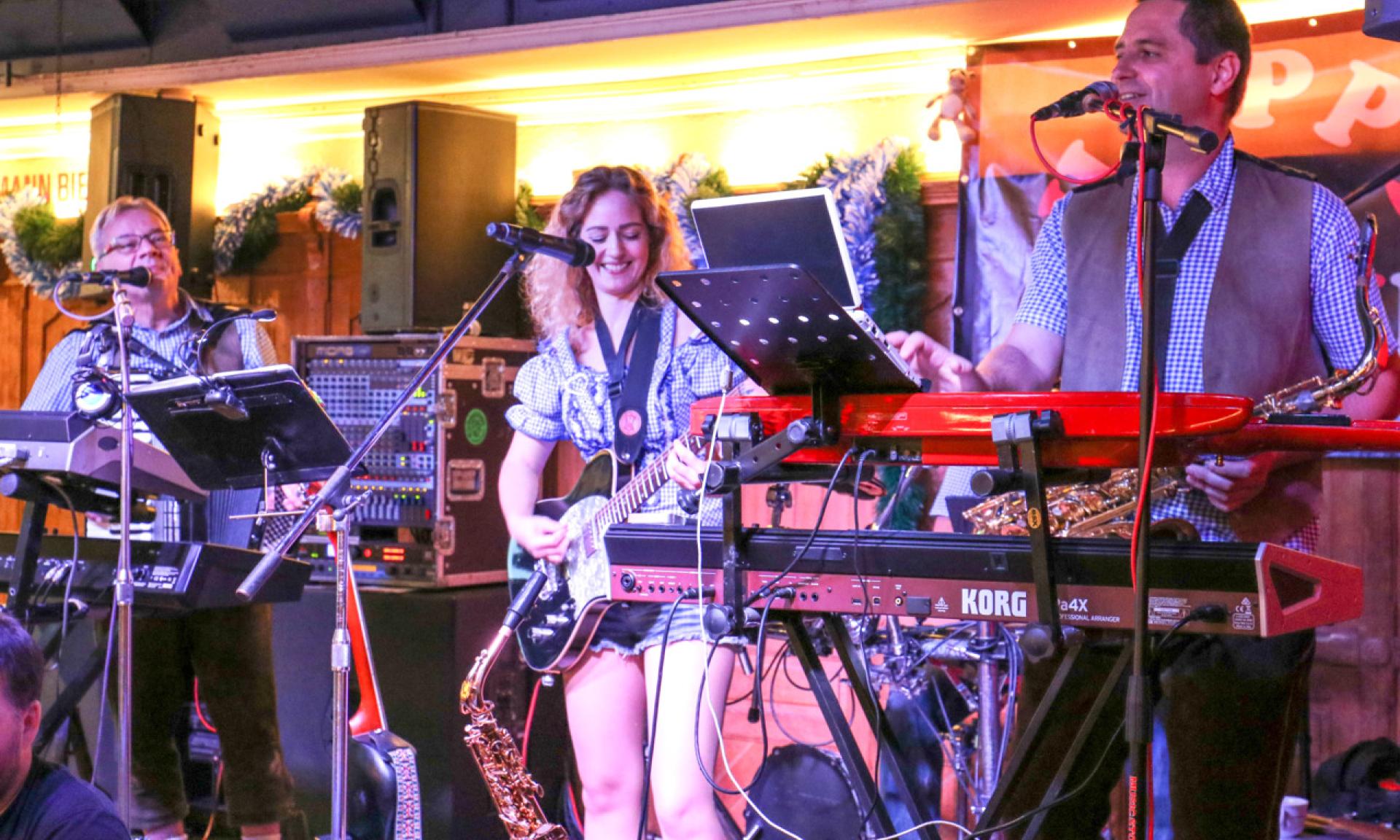 A vibrant band featuring a smiling female guitarist and two men on keyboards and saxophone performs lively music on a brightly lit stage in a festive Bierhalle.