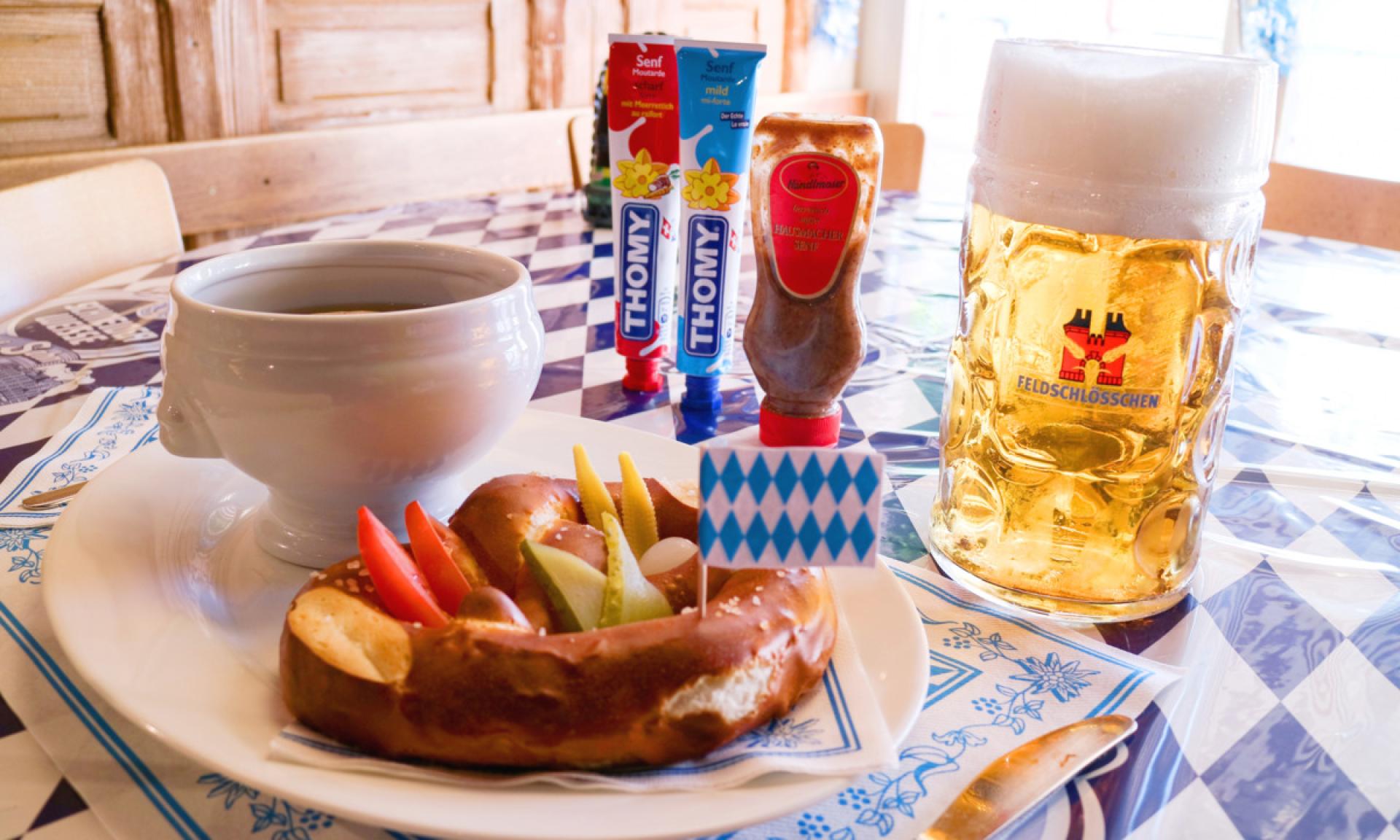 Traditional Bavarian meal featuring a Feldschlösschen beer mug, garnished pretzel, soup, and various mustards on a blue and white checkered tablecloth in a German beer hall setting.