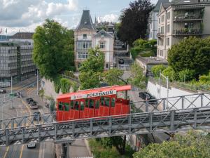 Vibrant red Polybahn funicular ascends its elevated track above a busy city street, surrounded by a mix of traditional and contemporary buildings and lush trees.