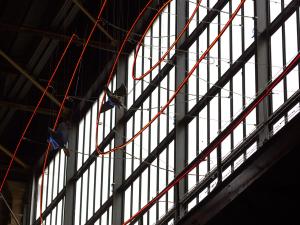 A vast industrial space showcases Niki de Saint Phalle's L'ange protecteur art installation, featuring dark abstract figures suspended amidst a stark metal beam grid, illuminated by striking red neon lines.