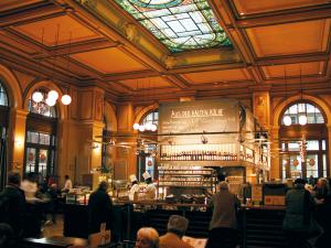A warm, bustling grand cafe interior features an elaborate stained-glass skylight, rich wooden coffered ceiling and paneling, and patrons gathered around tables and a central bar.