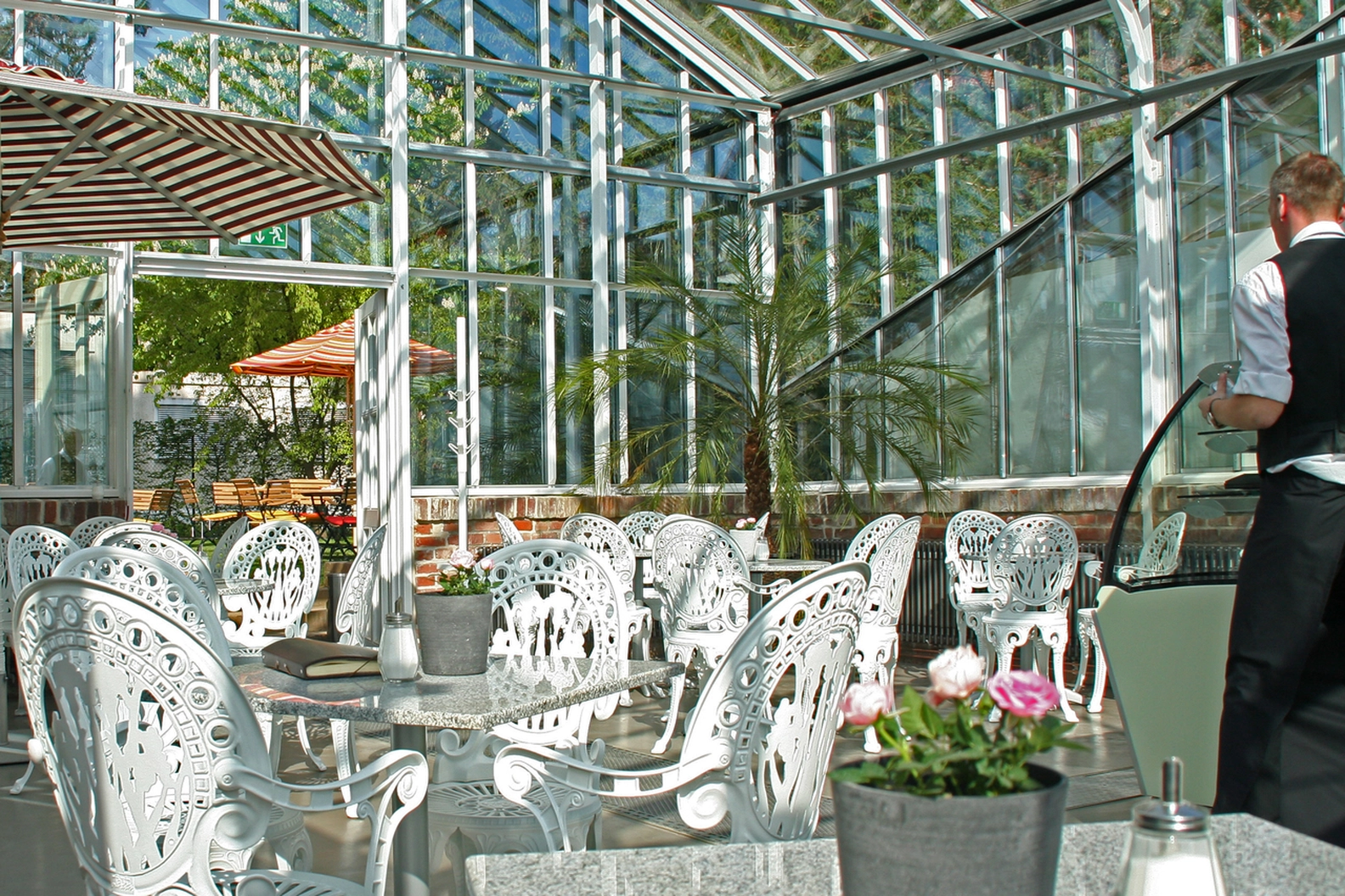 Sunlit glass conservatory cafe with elegant white cast iron tables and chairs, tropical plants, and a charming outdoor patio visible, showcasing a perfect cafe in the sunshine.