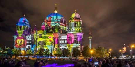 Berliner Dom illuminated by vibrant, colorful light projections during the Berlin Festival of Lights, with the Fernsehturm visible in the background.