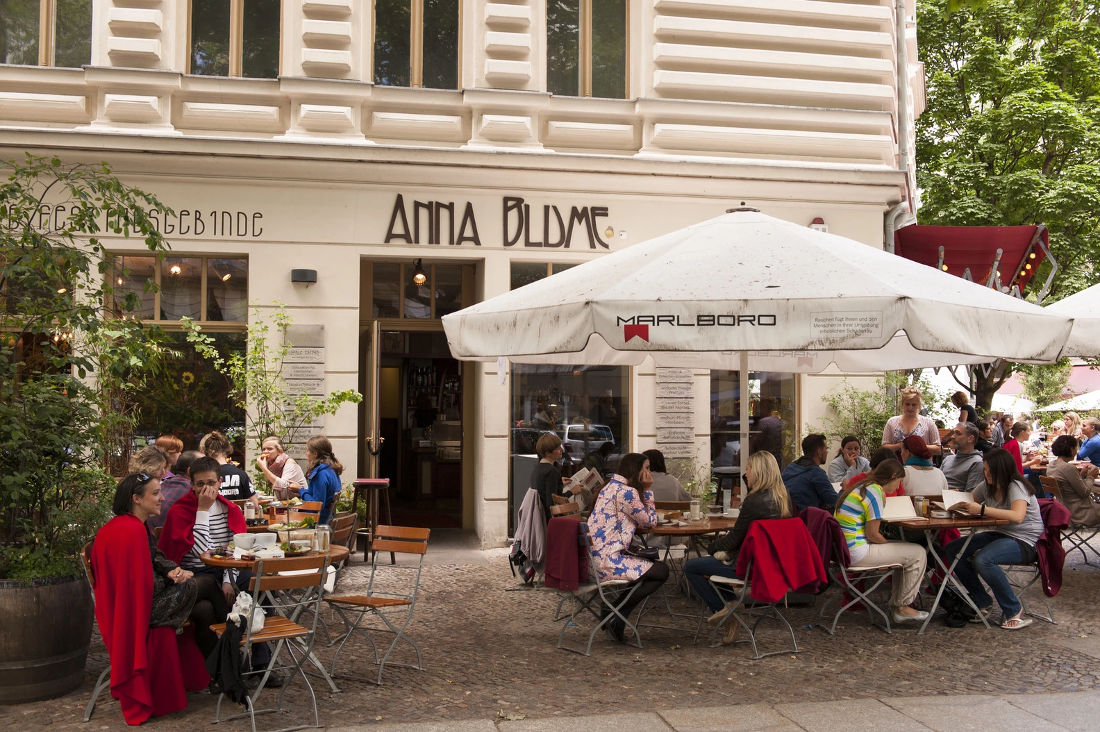 Anna Blume cafe's sunny outdoor patio, bustling with customers enjoying homemade cakes and drinks at tables under large white umbrellas.