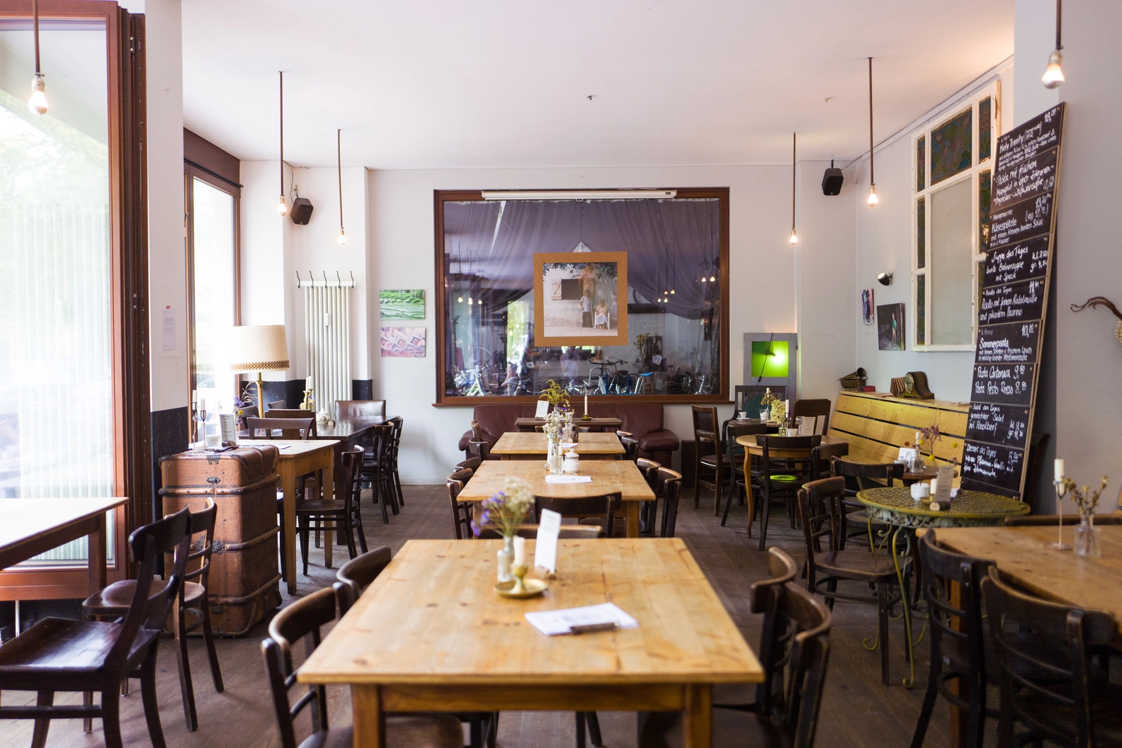 Bright, inviting interior of Kaffee A Horn cafe featuring rows of wooden tables, dark chairs, unique vintage decor including a large mirror and chalkboard menu, creating a welcoming and cosy atmosphere.
