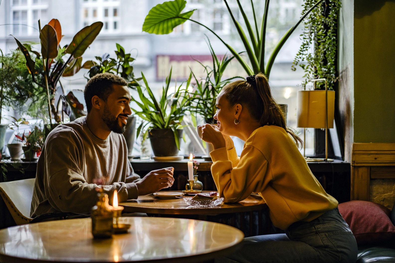 Smiling couple enjoying coffee and conversation at a cozy, plant-filled book canteen cafe, illuminated by natural light and warm candles.