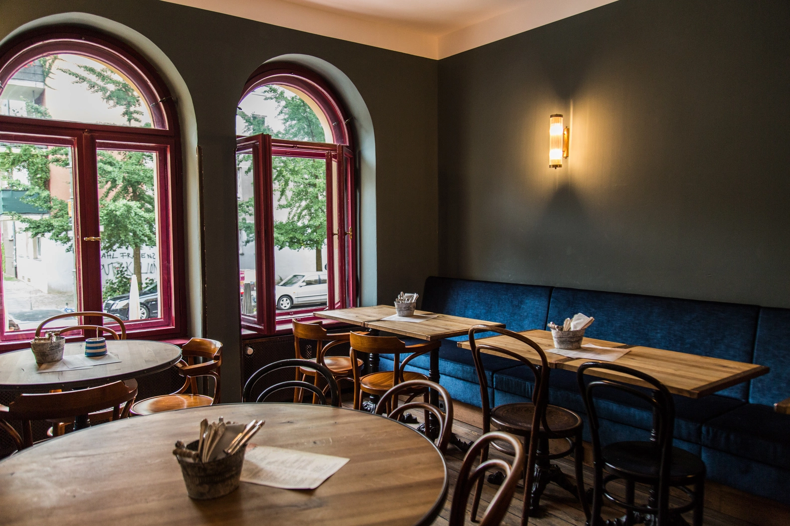 Betty N Caty cafe interior with arched red windows, blue banquette seating, and wooden tables, bathed in natural light overlooking the neighborhood street.