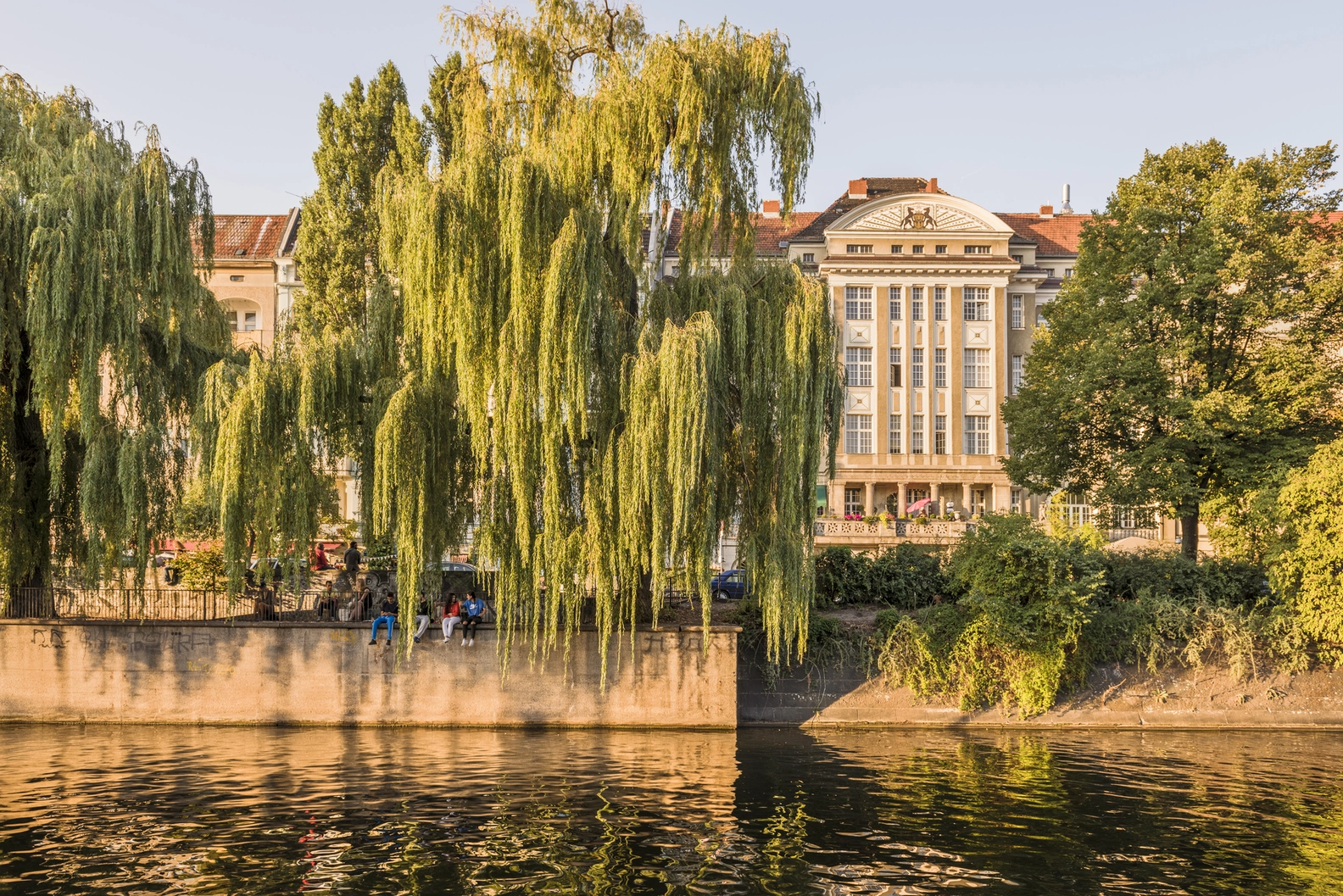 Sunny Berlin riverside with a grand historic building, weeping willows, and people enjoying the warm outdoor cafe ambiance.