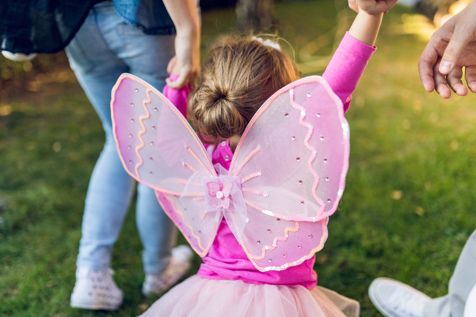 Young child in vibrant pink fairy costume with iridescent wings, held by adults at Britzer Gartennacht in Berlin, capturing the magical summer night atmosphere.