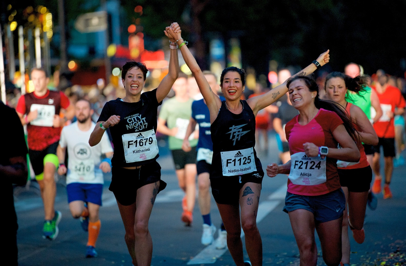 Triumphant women runners Katharina and Nadin-Isabell celebrate with arms raised during the Adidas City Night 2017 race in Berlin, embodying summer night sporting heroism.