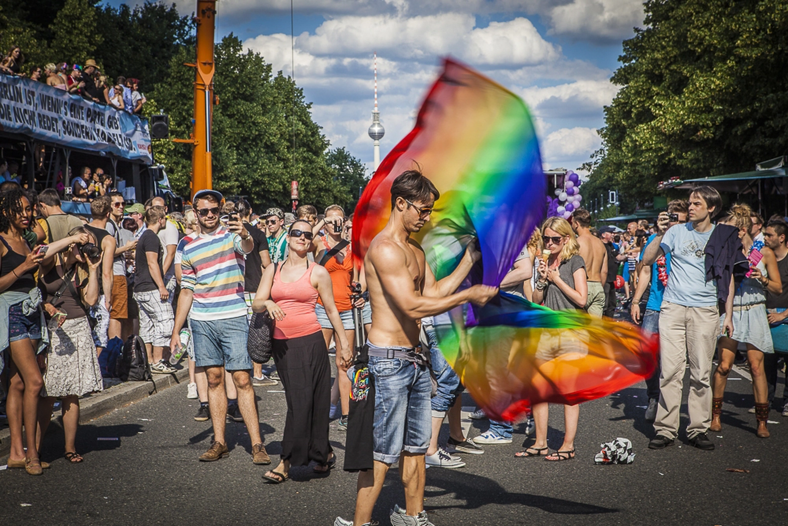 A shirtless man proudly waves a large rainbow Pride flag at a vibrant Berlin Pride Month celebration, surrounded by a festive crowd and the iconic TV Tower under a summer sky.