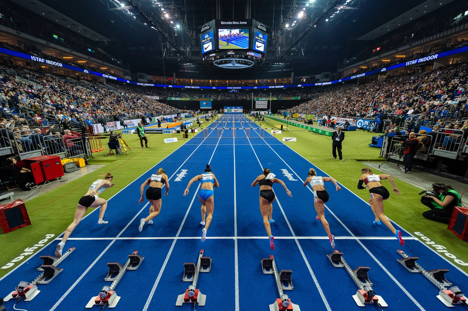Elite female athletes burst from starting blocks on a blue track during an indoor athletics race at ISTAF in Berlin's Mercedes-Benz Arena, surrounded by cheering fans.