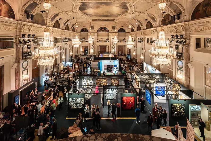 Grand art exhibition in an ornate hall with sparkling chandeliers, showcasing diverse contemporary artworks as visitors browse elaborate displays during an autumn design district art event.