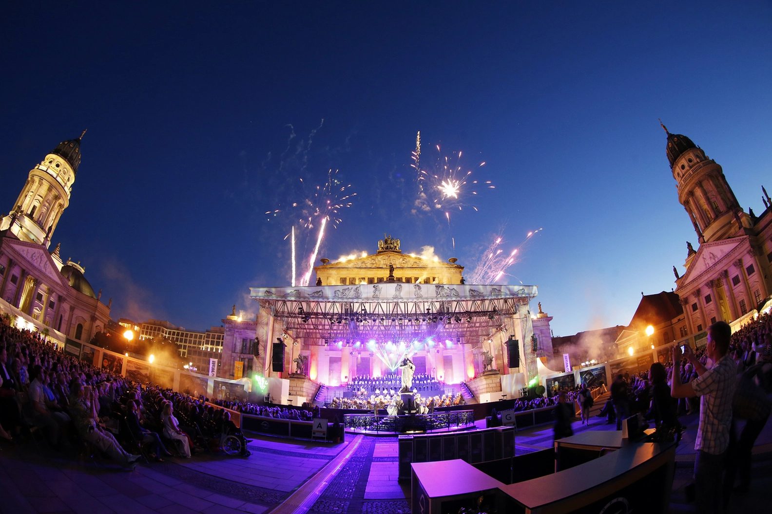 Gendarmenmarkt in Berlin hosts a vibrant open-air classical music concert with fireworks illuminating the sky above the Konzerthaus and an audience enjoying the summer night.