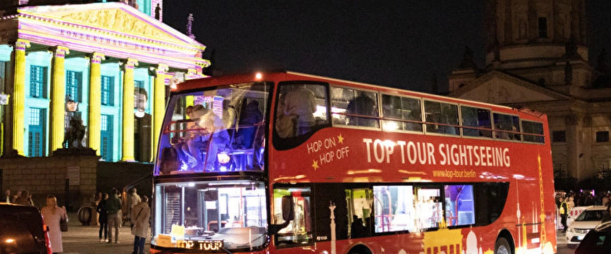Red double-decker sightseeing bus with tourists on a night tour, passing the illuminated Konzerthaus during the Berlin Festival of Lights.