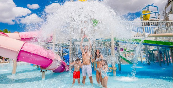 Family cooling off under a large water splash at a Madrid water park, a refreshing activity for August.