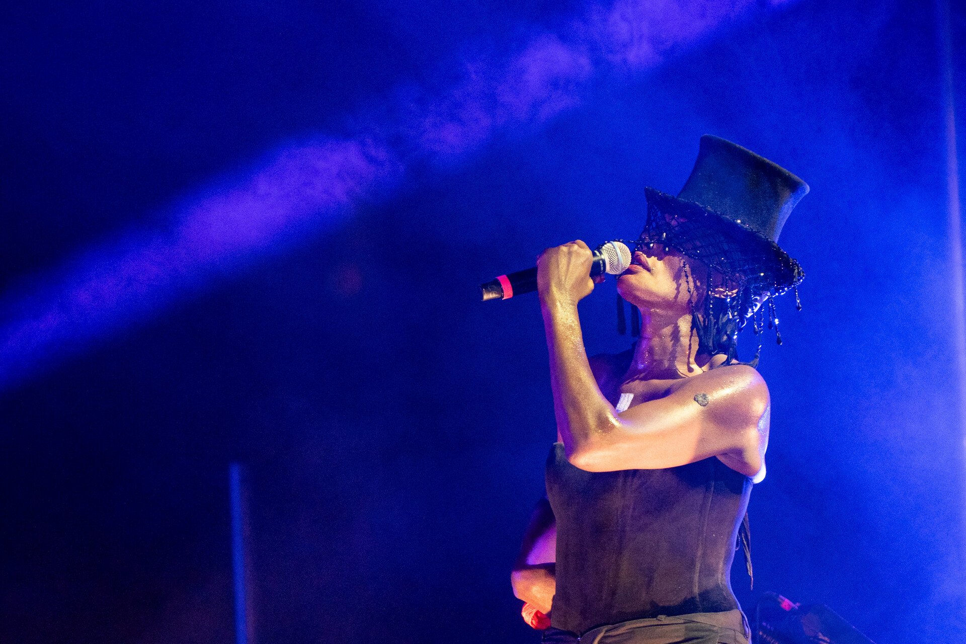 Grace Jones singing into a microphone on stage, wearing a black top hat, under vibrant blue and purple concert lighting.
