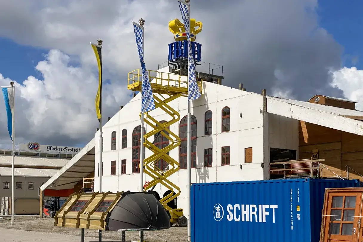 Oktoberfest Baustelle featuring the Hacker-Festzelt and Hofbräuhaus tent with its crown logo under construction, alongside a yellow scissor lift and Bavarian flags.