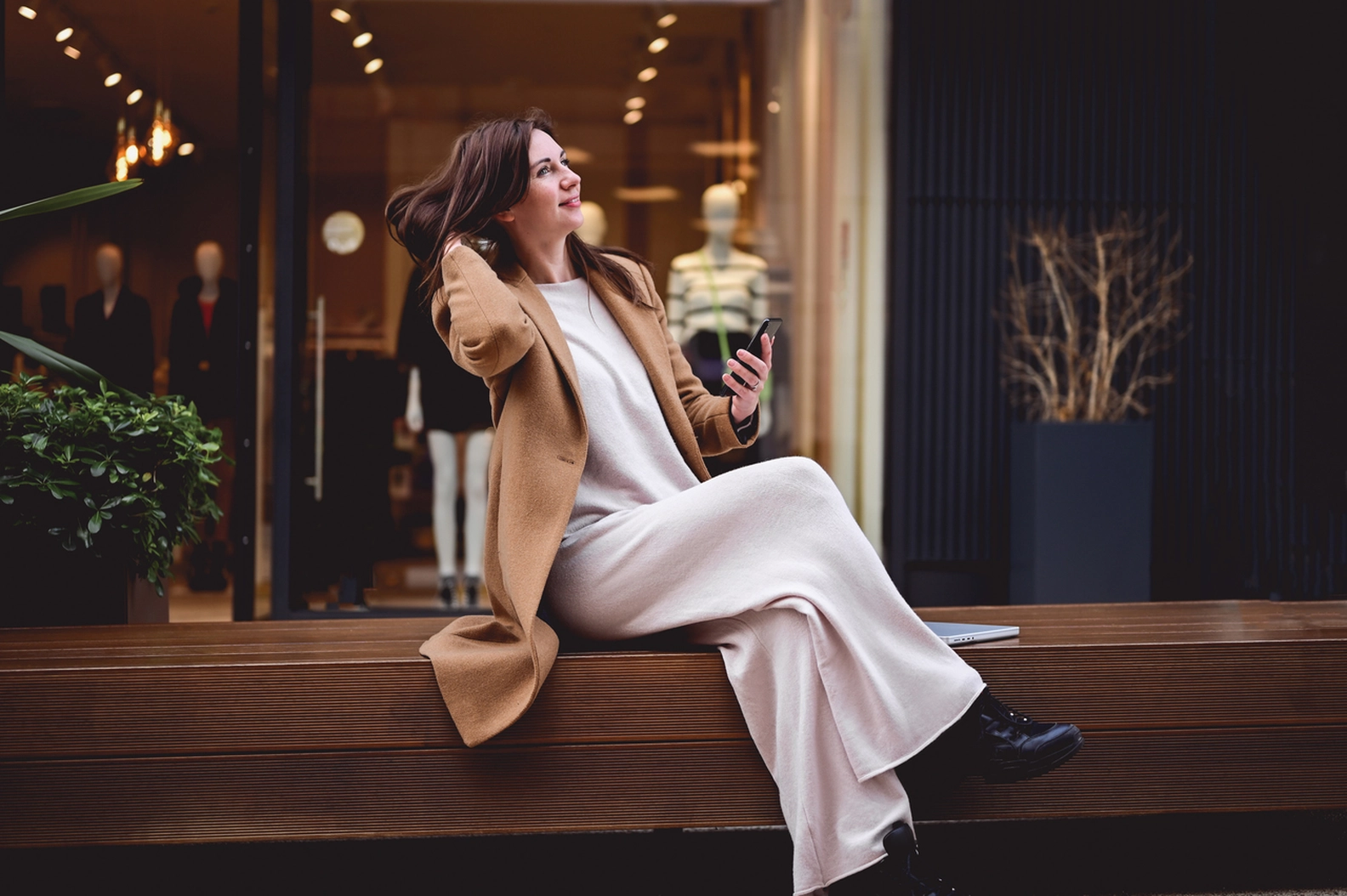 Stylish woman in a camel coat and flowing wide-leg outfit relaxing on a bench, showcasing current fashion trends during Berlin Fashion Week.
