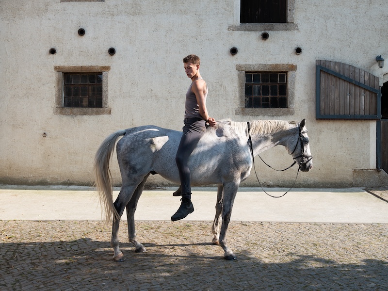 Young man riding a dapple grey horse bareback in front of a rustic building, for Galerie Nord Kunstverein Tiergarten exhibition.