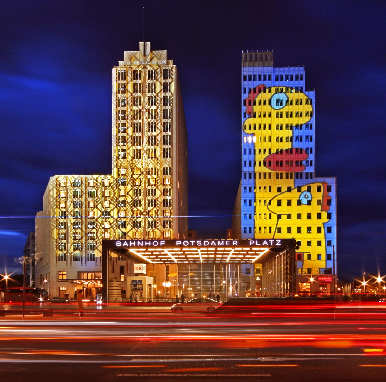 Potsdamer Platz buildings illuminated during Berlin Festival of Lights, featuring a golden geometric light projection on one tower and a colorful character projection on another, with the Bahnhof Potsdamer Platz station sign and blurred traffic light trails.