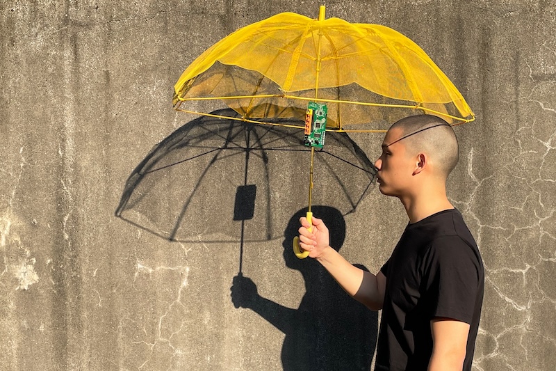 Person holds a deconstructed yellow umbrella featuring a prominent circuit board, casting a strong shadow on a concrete wall. This unique art piece is part of an exhibition at Kunstquartier Bethanien for The Week August 4-24, 2025.