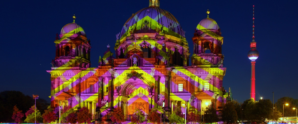 Berliner Dom illuminated with vibrant purple and yellow light art during Berlin's Festival of Lights, featuring the iconic red Berlin TV Tower in the background. A highlight for any lightseeing walking tour.