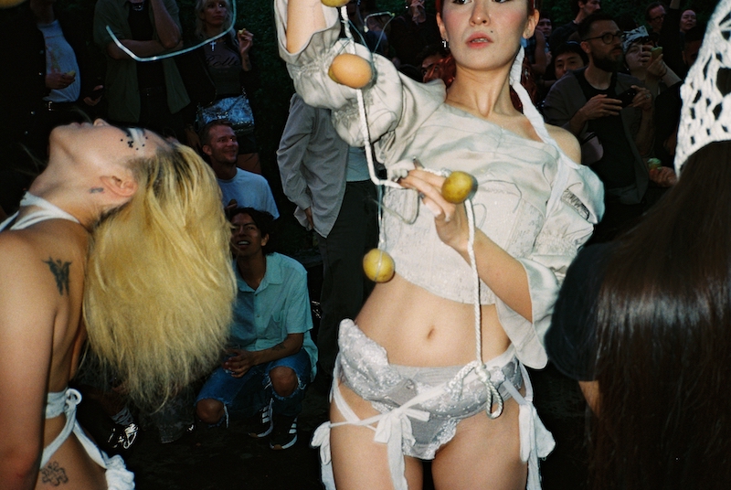 Caique Tizzi and YBDG performers at Schinkel Pavillon's 'Feeding Frenzy'. A performer in avant-garde attire offers lemons on a string for a performance-initiation-drinks ritual, as another participant's head is thrown back.