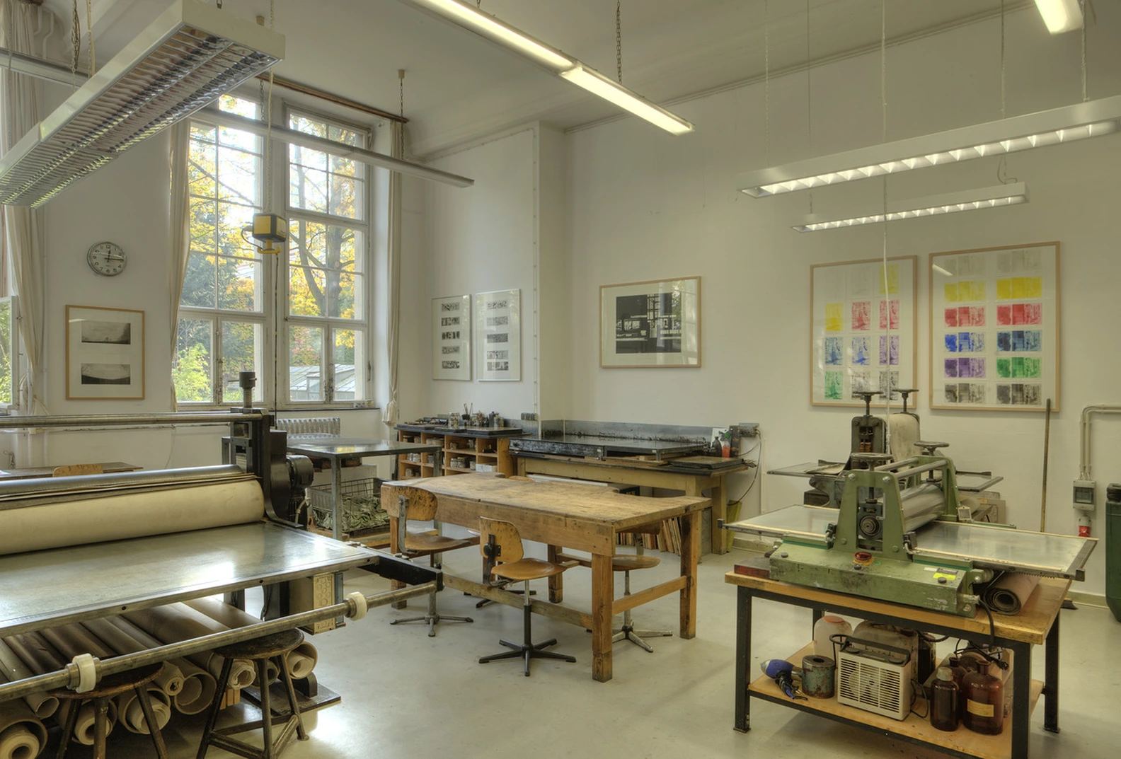 Spacious, well-lit printmaking studio at Berlin University of the Arts, featuring traditional presses, workbenches, large windows, and vibrant abstract art prints displayed on white walls.